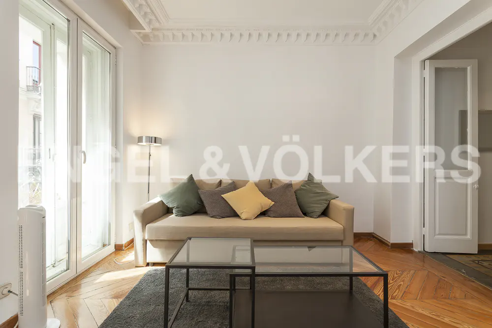 Living room with beige sofa, colorful pillows, glass coffee tables, and hardwood floors. Balcony doors let in natural light.