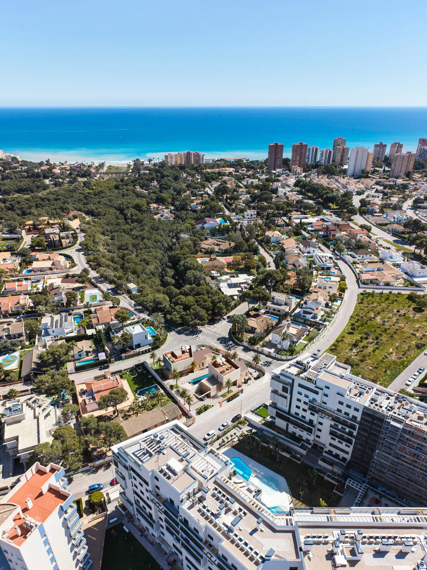 Aerial view of a coastal town with white buildings, green trees, and a blue ocean under a clear sky.