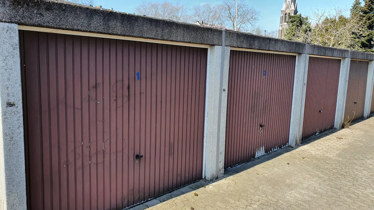 Row of brown garage doors with concrete frames, numbered 1 and 2, on a paved surface. A church steeple is visible in the background.
