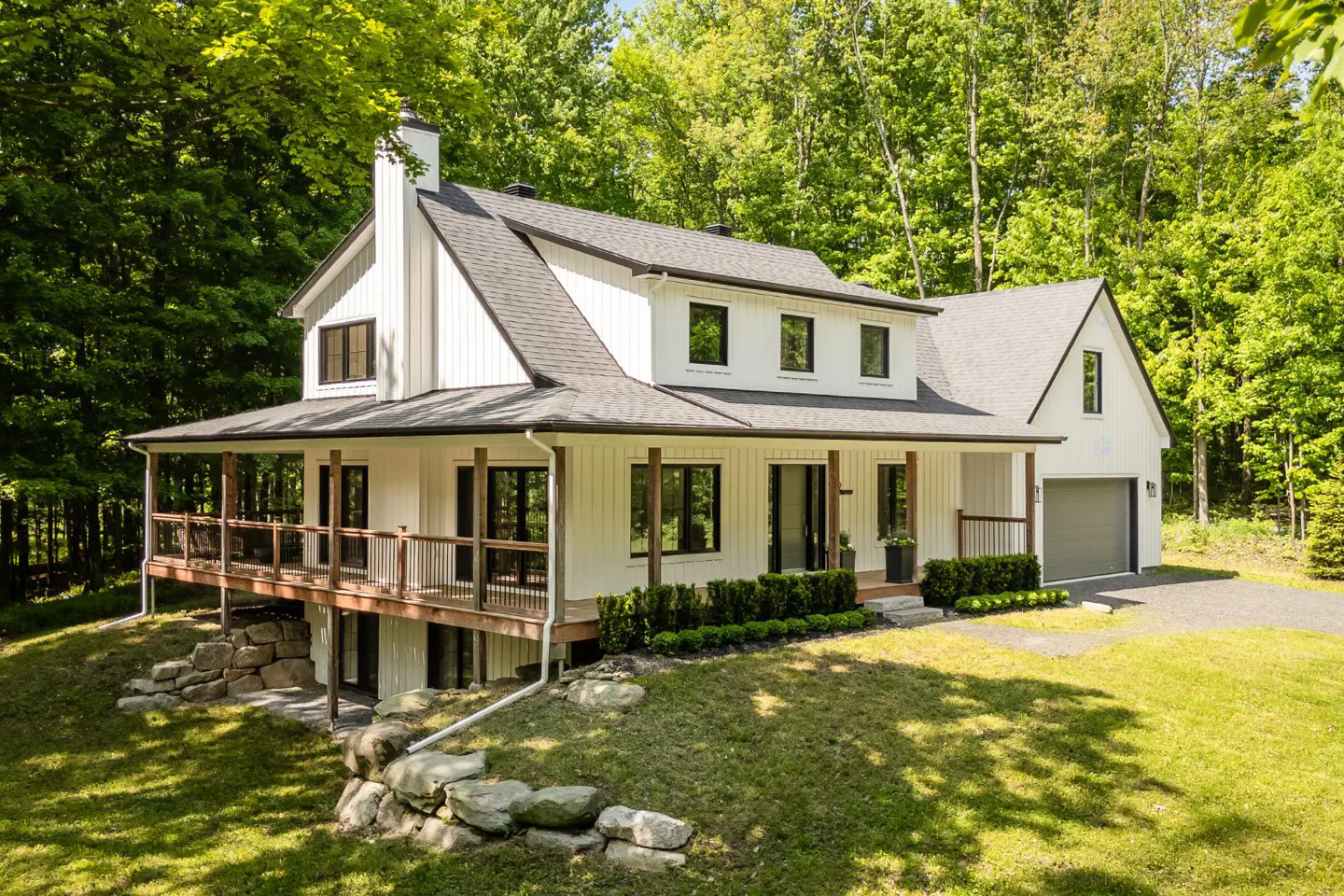 A modern farmhouse with white siding, black trim, and a gray roof sits on a green lawn surrounded by trees.