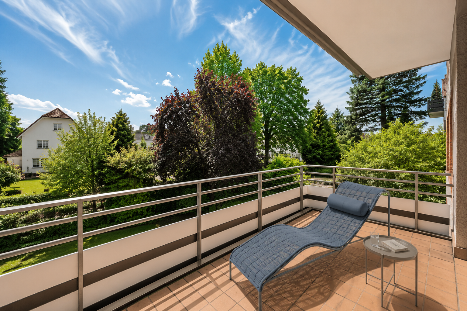 A balcony with a blue lounge chair and small table, overlooking a lush green garden and a white house under a sunny sky.
