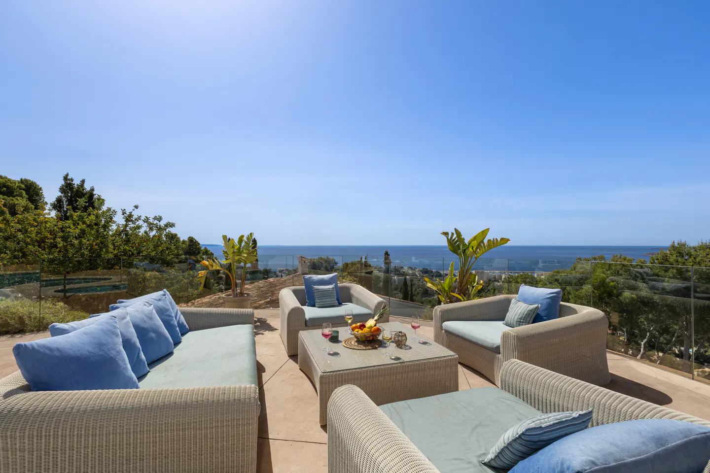 Outdoor patio with wicker furniture, blue cushions, and a glass table with fruit and wine. Ocean view and clear blue sky in the background.