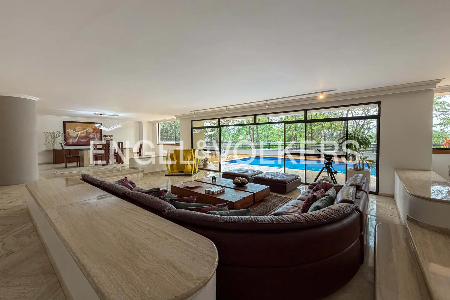 A spacious living room with a brown leather sofa, a wooden coffee table, and a view of a blue swimming pool through large windows.