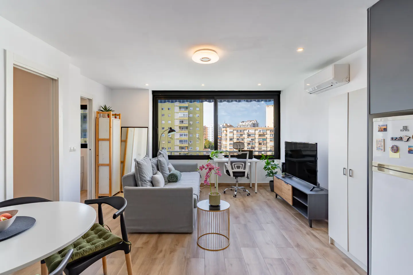 Bright living room with a gray sofa, white desk, and large window overlooking city buildings. Wood floors and modern decor.
