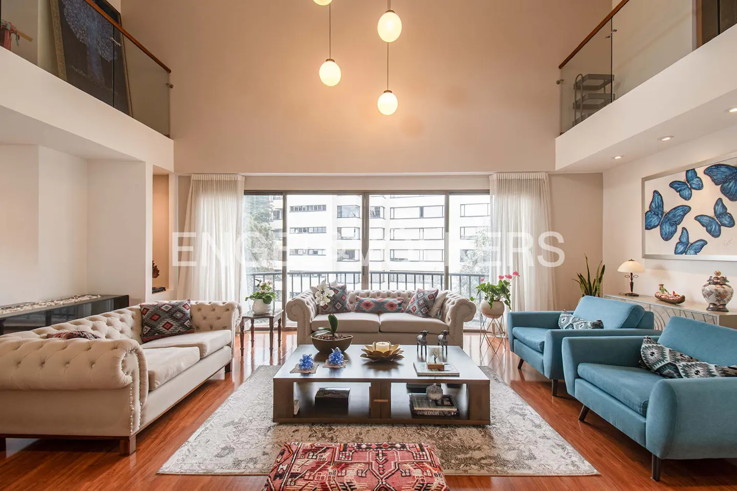 Bright living room with beige sofas, blue chairs, and a large window overlooking a city. A modern chandelier hangs above a coffee table.