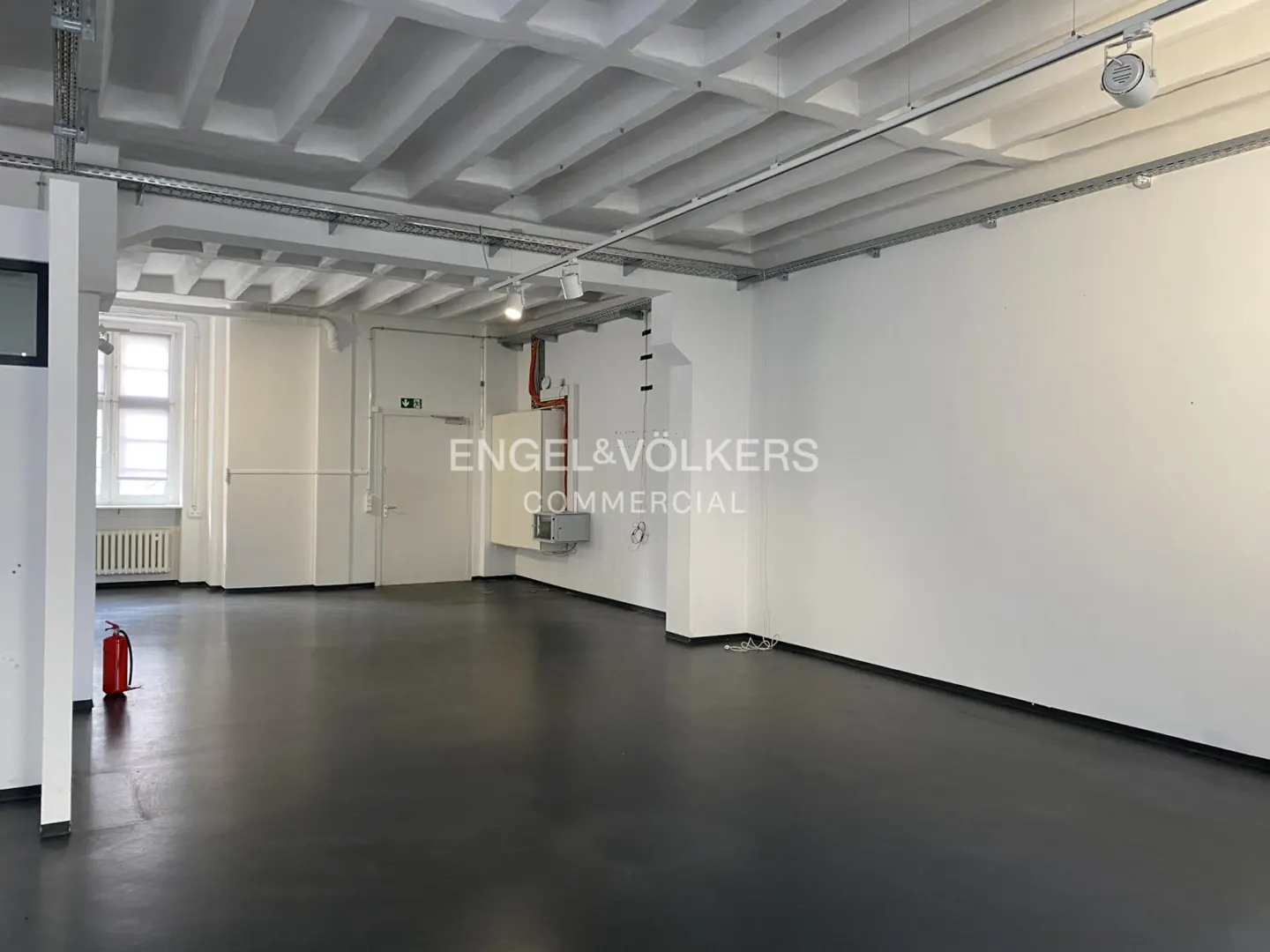 Empty commercial space with white walls, dark grey flooring, exposed ceiling beams, and track lighting.