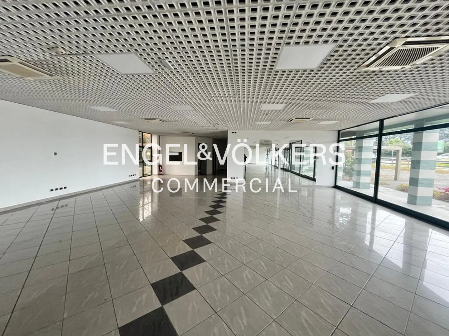 Empty commercial space with white walls, tiled floor with black squares, and a lattice ceiling. Engel & Völkers logo is visible.