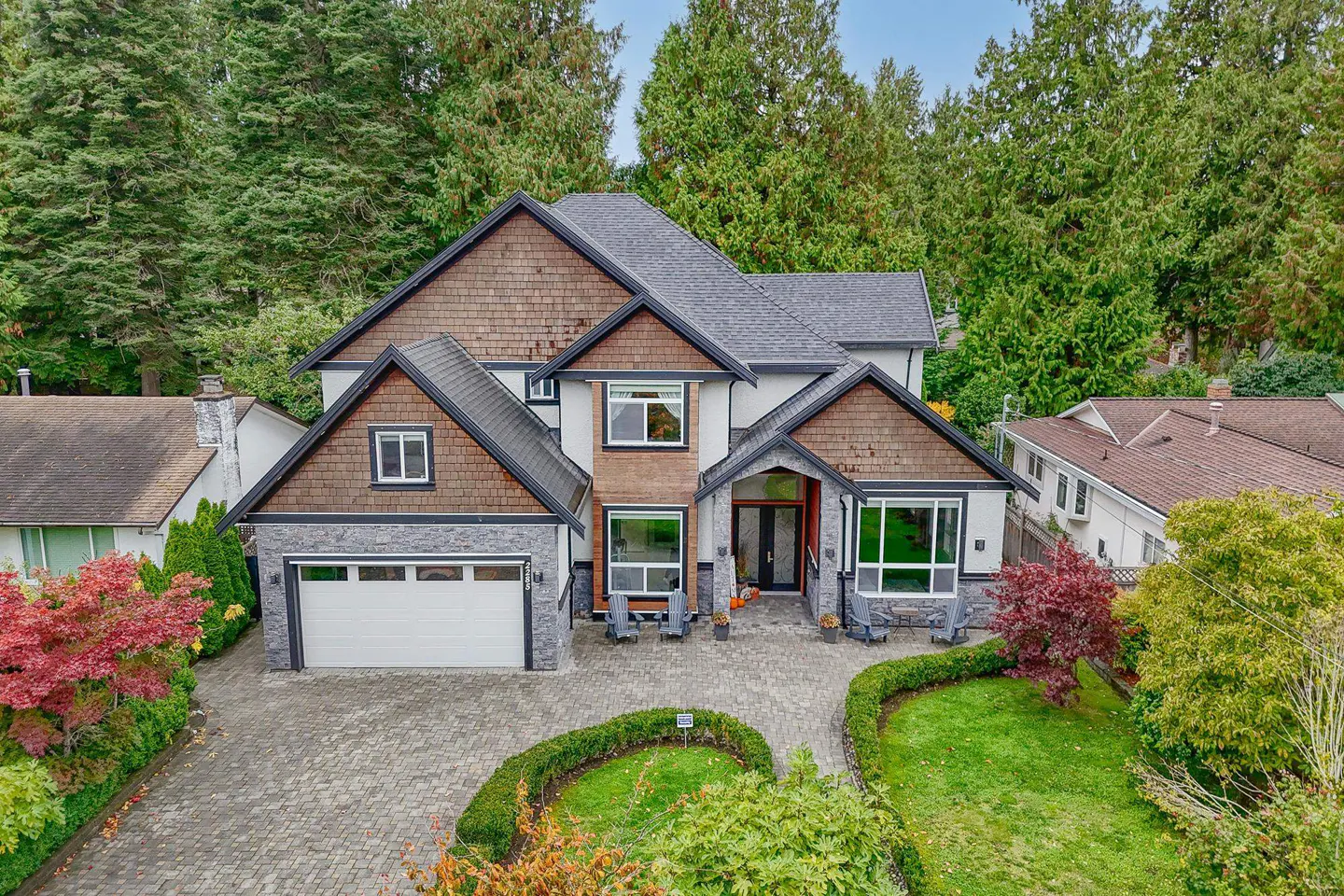 Two-story house with gray roof, brown shingles, stone accents, and white garage door. Green trees in background.