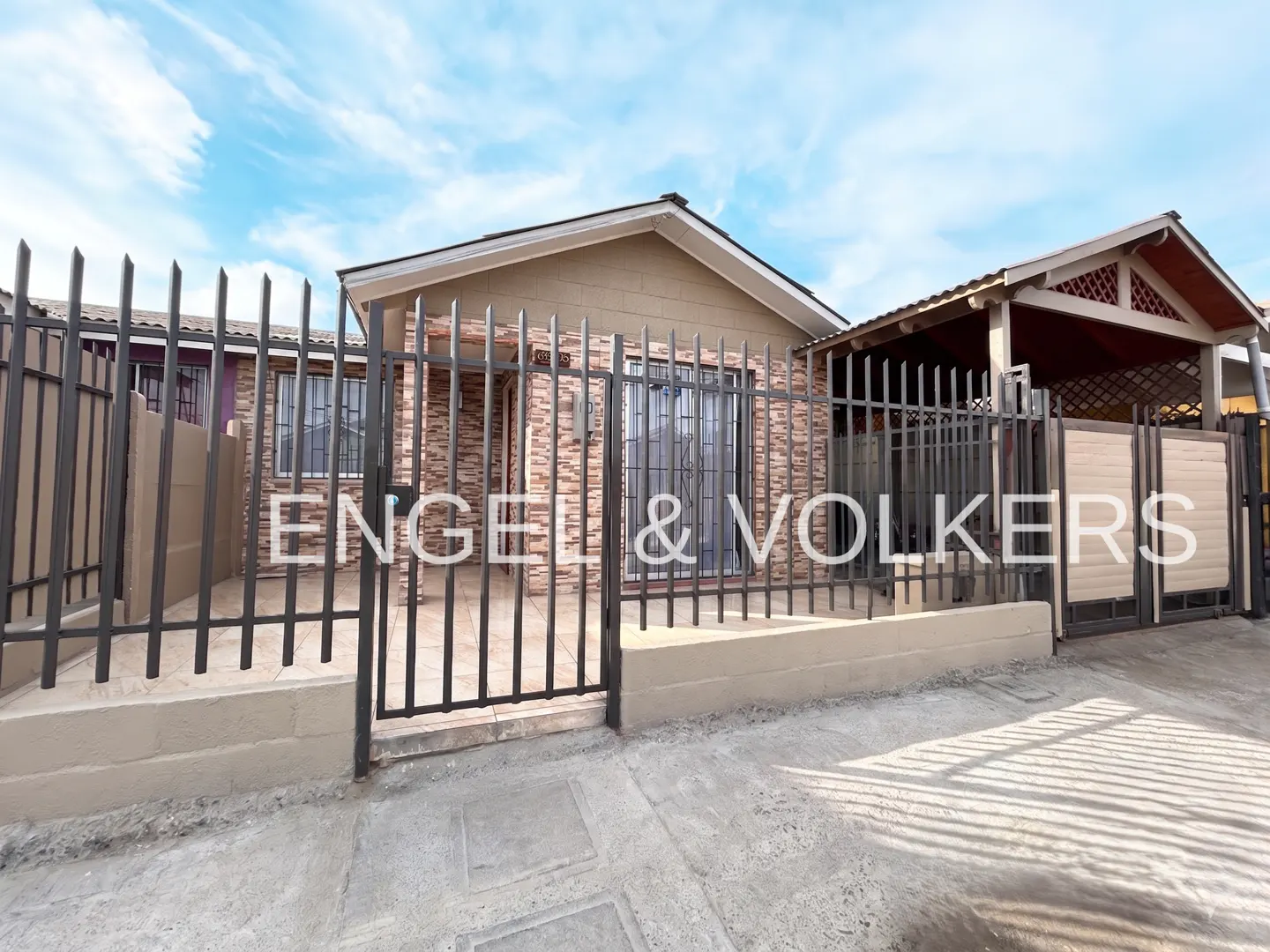 Single-story house with stone-textured facade, metal security fence, and a covered carport under a blue sky.