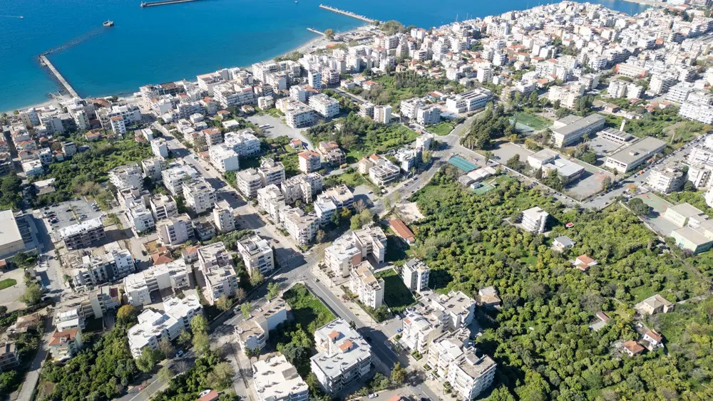 Aerial view of a coastal city with white buildings, green trees, and blue sea. A pier extends into the water.