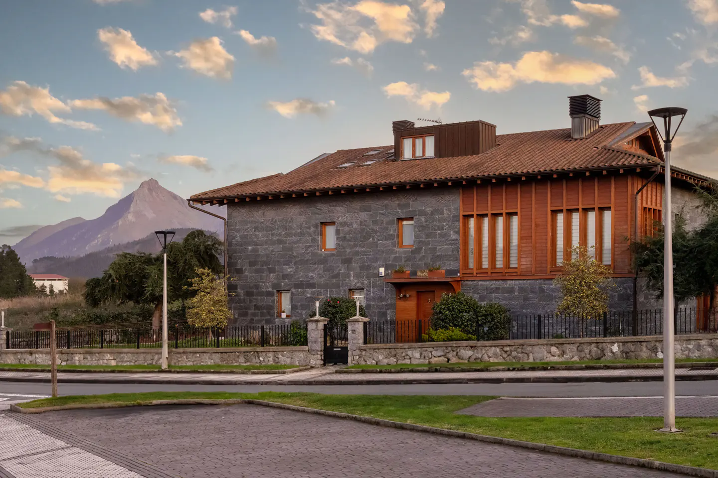 A two-story house with gray stone and wood siding, a brown tile roof, and a mountain backdrop.