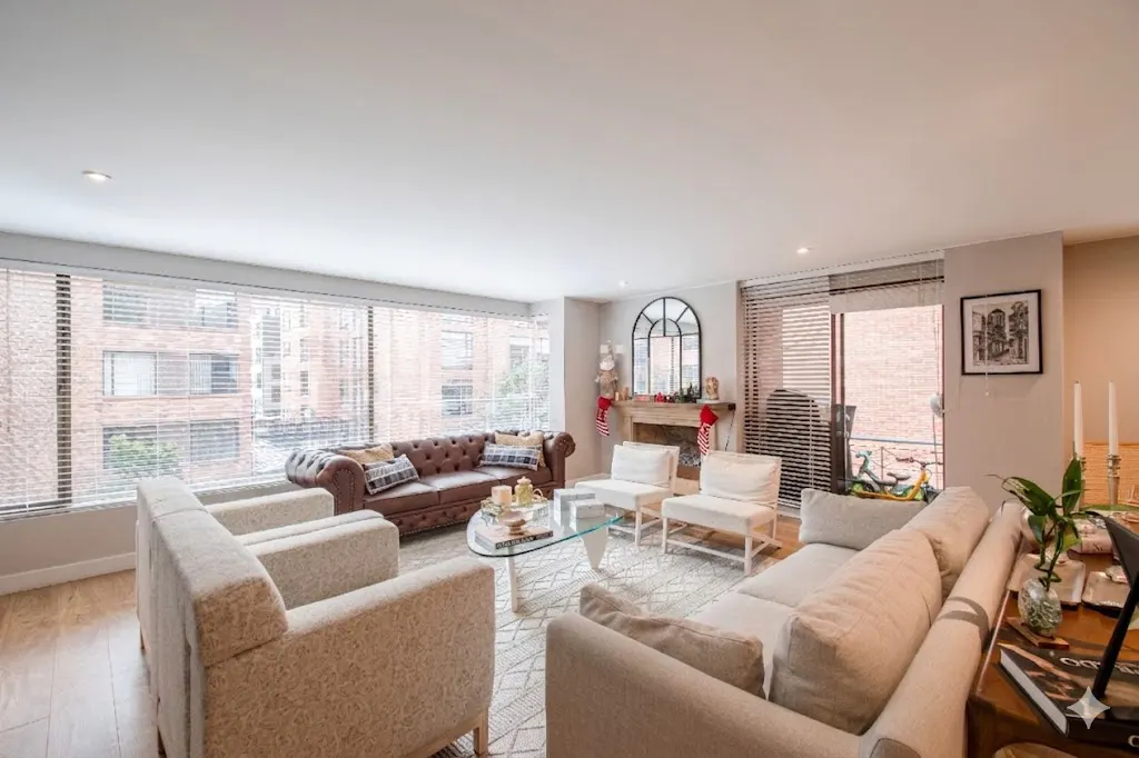 Bright living room with brown leather sofa, white chairs, glass table, and fireplace with Christmas stockings. Large windows with blinds.