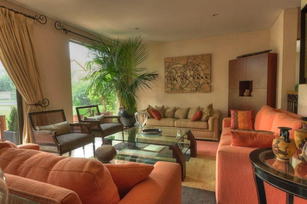 A living room with orange and beige sofas, a glass coffee table, and two armchairs by a large window. A tall plant and artwork add to the room's decor.
