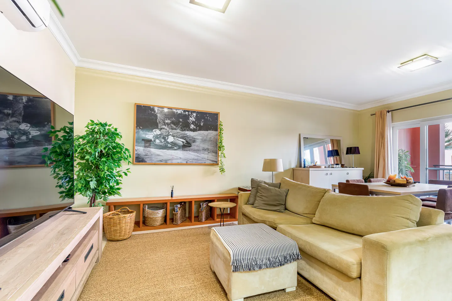 Living room with beige walls, a tan sectional sofa, and a jute rug. A black and white photo hangs above a wooden shelf.