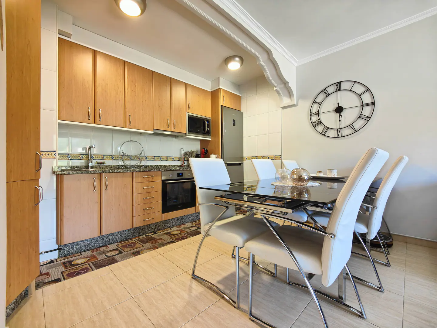 A kitchen and dining area with light wood cabinets, granite countertops, and a black glass table with white chairs. A large clock hangs on the wall.