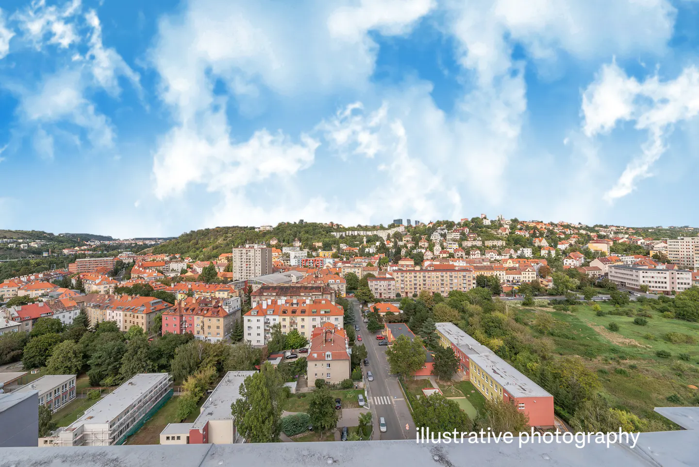 Cityscape view with red-roofed buildings, green trees, and a blue sky with white clouds. A road runs through the center of the scene.
