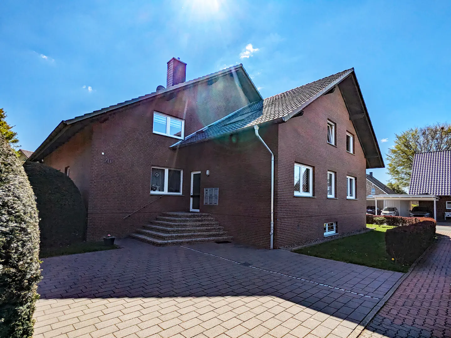 Two-story brick house with a chimney, white-framed windows, and a paved driveway under a sunny blue sky.