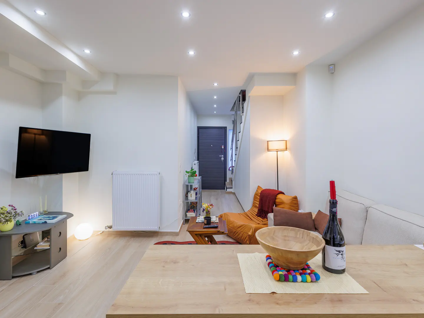 Bright living room with a TV, radiator, and table with a wine bottle. A hallway leads to a door and stairs.