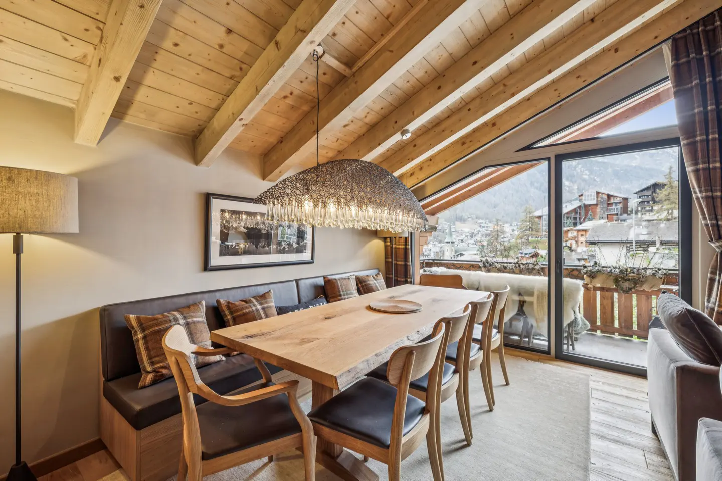 Dining area with wood table, chairs, and bench seating. A large window shows a mountain town view. Wooden ceiling beams add rustic charm.