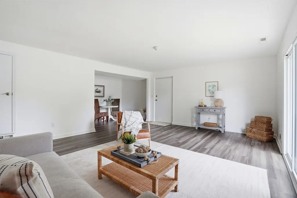 Bright living room with white walls, gray wood floors, and a woven coffee table on a white rug. Dining room visible through an archway.