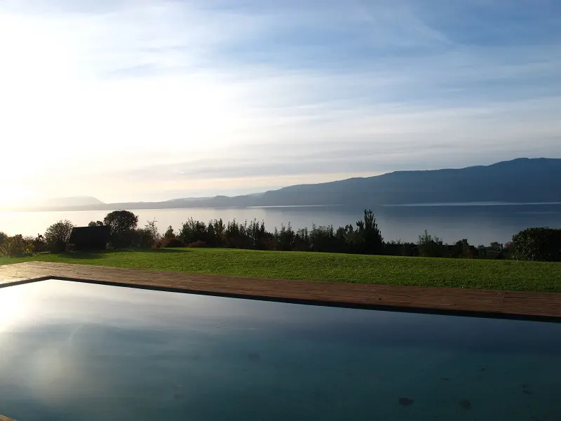 Scenic view of a pool reflecting the sky, with a green lawn, trees, and a lake with mountains in the background.