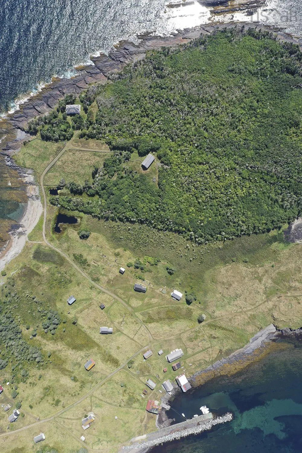 Aerial view of a green island with buildings, paths, and a rocky coastline meeting the blue ocean.