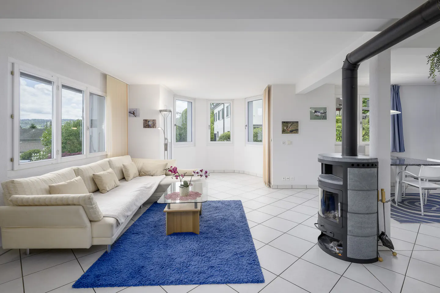 Bright living room with white walls, tiled floor, and large windows. A cream sofa sits on a blue rug, with a modern fireplace in the corner.