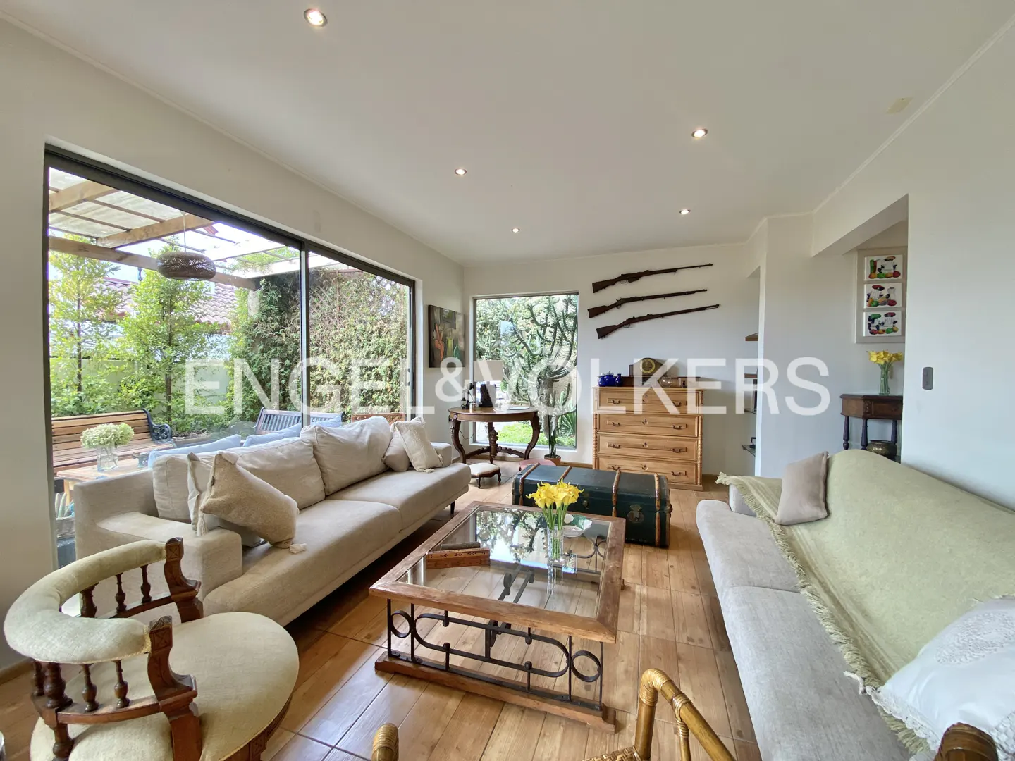 A bright living room with sofas, a glass coffee table, and wooden floors. Large windows offer a view of the garden. Three rifles are mounted on the wall.