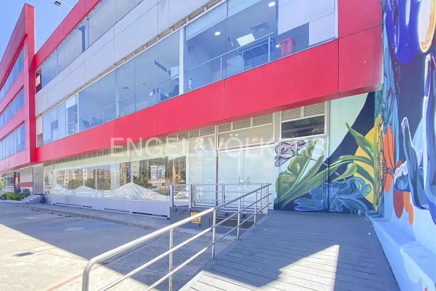 Exterior view of a modern, multi-story Engel & Völkers office building with red accents, large windows, and a colorful mural. A ramp with railings leads to the entrance.
