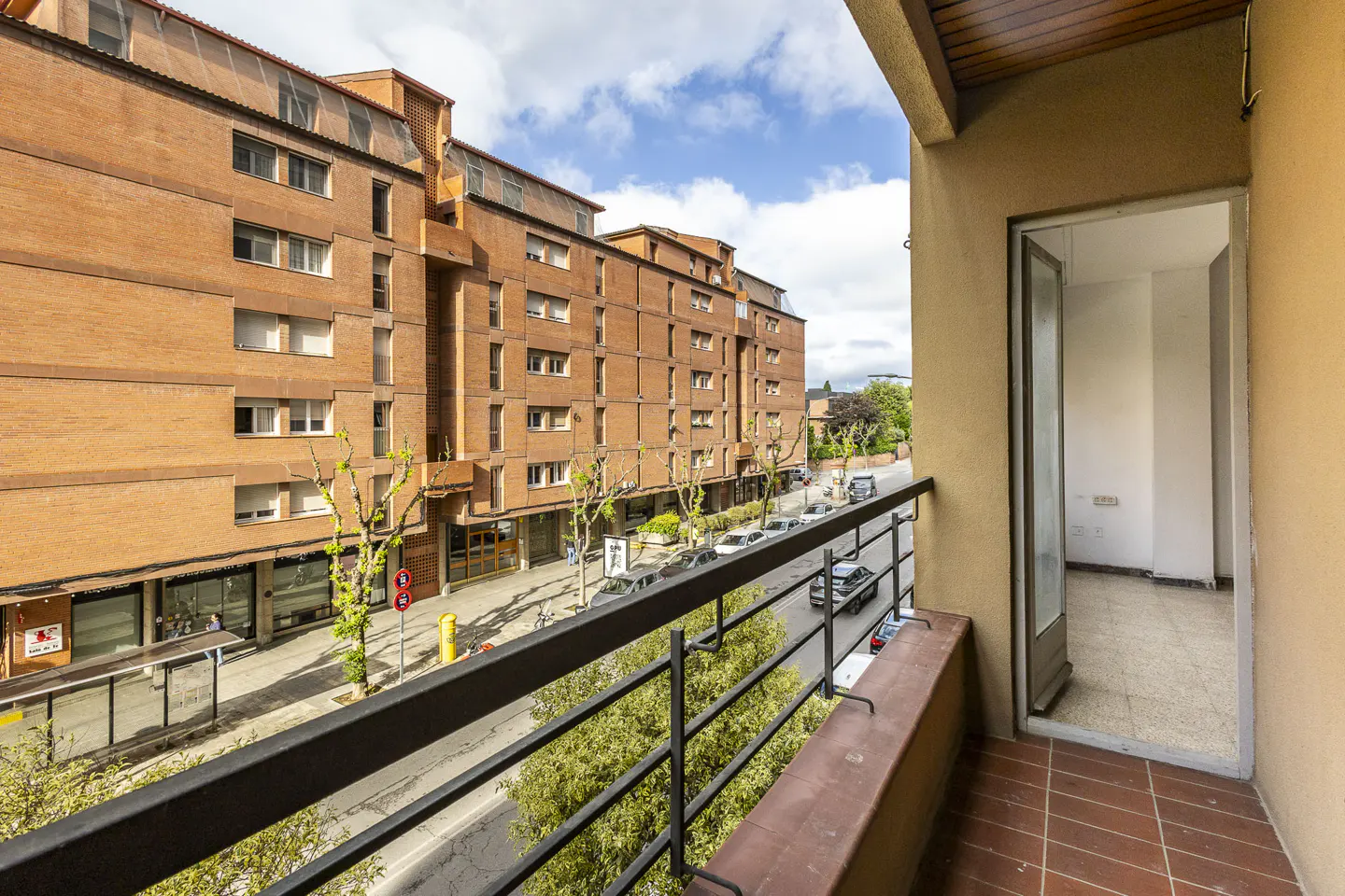 View from a balcony with black railings overlooking a street with brick buildings and cars. An open door leads to an empty room.