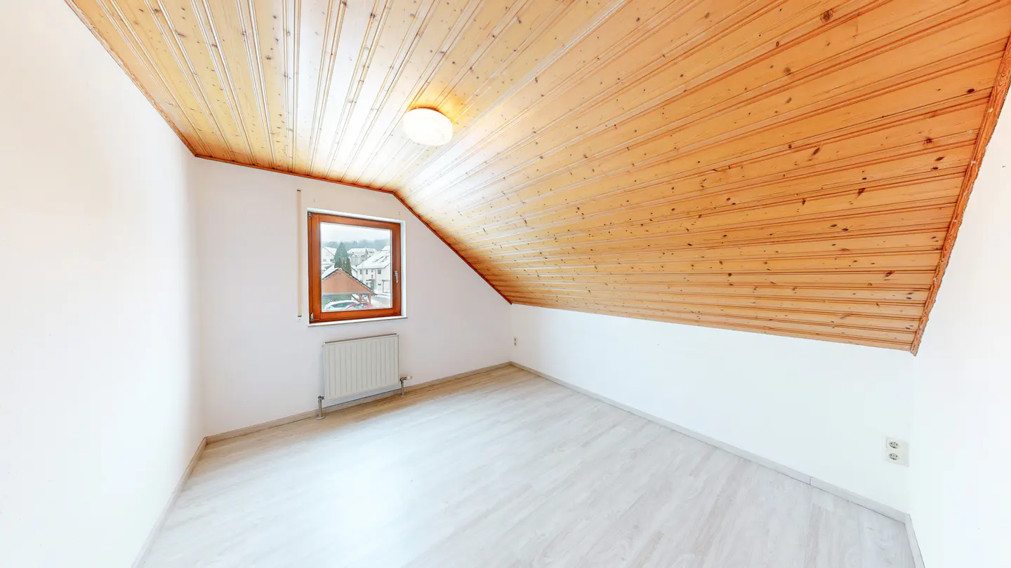 Empty attic room with white walls, light wood flooring, and a sloped wooden ceiling with a central light fixture.