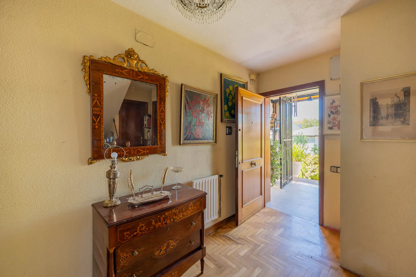 Foyer with a wooden dresser, ornate mirror, and open door leading to a bright outdoor area. Art hangs on the cream-colored walls.