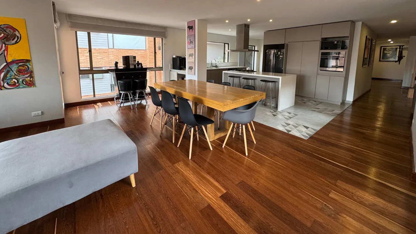 Open-plan living area with hardwood floors, a wooden dining table, grey chairs, and a modern kitchen with a breakfast bar.