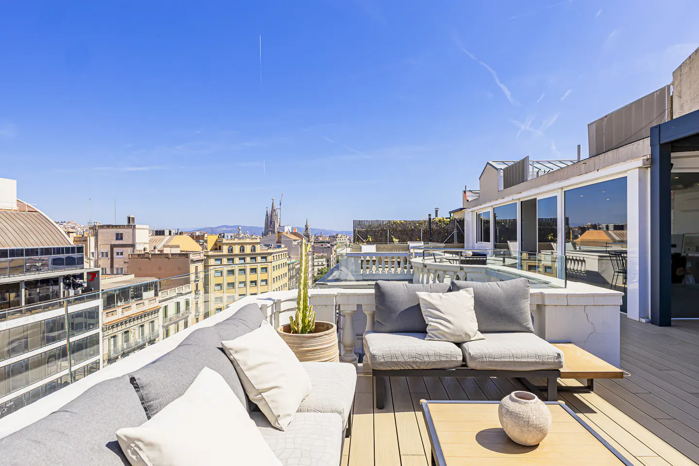 Rooftop patio with gray sofas, white pillows, and a wooden table. Barcelona cityscape with Sagrada Familia in the background under a clear blue sky.
