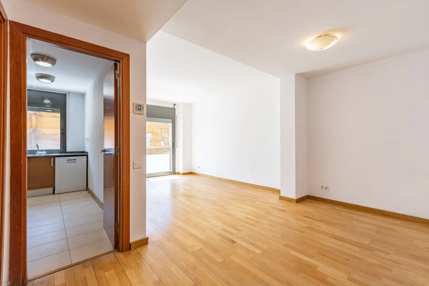 Bright, empty room with wood floors and white walls. A kitchen is visible through a doorway on the left. A glass door leads outside.