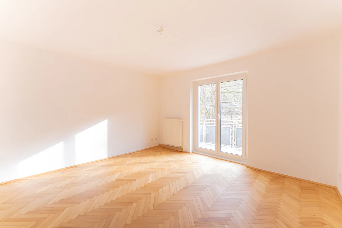 Bright, empty room with herringbone wood floors, white walls, and a glass door to a balcony.
