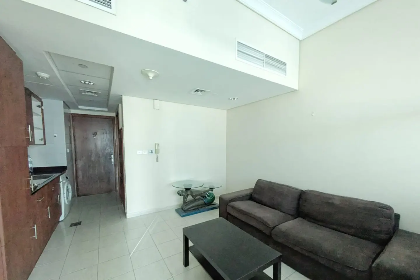 A living room with a dark brown sofa, black coffee table, and a small glass table with a decorative base near a kitchen.