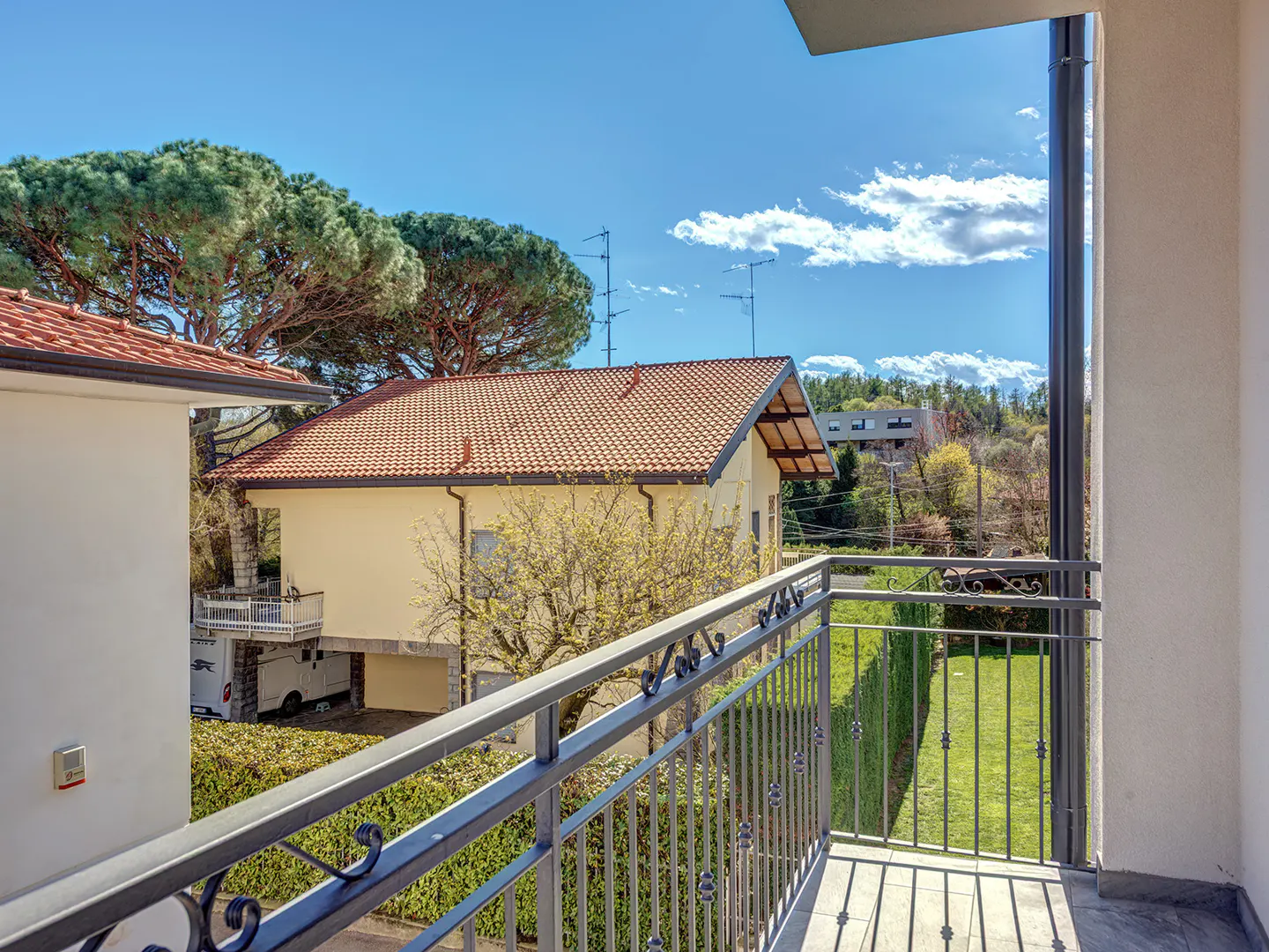 View from a balcony with metal railings overlooking a yard, houses with orange tile roofs, and trees under a blue sky.