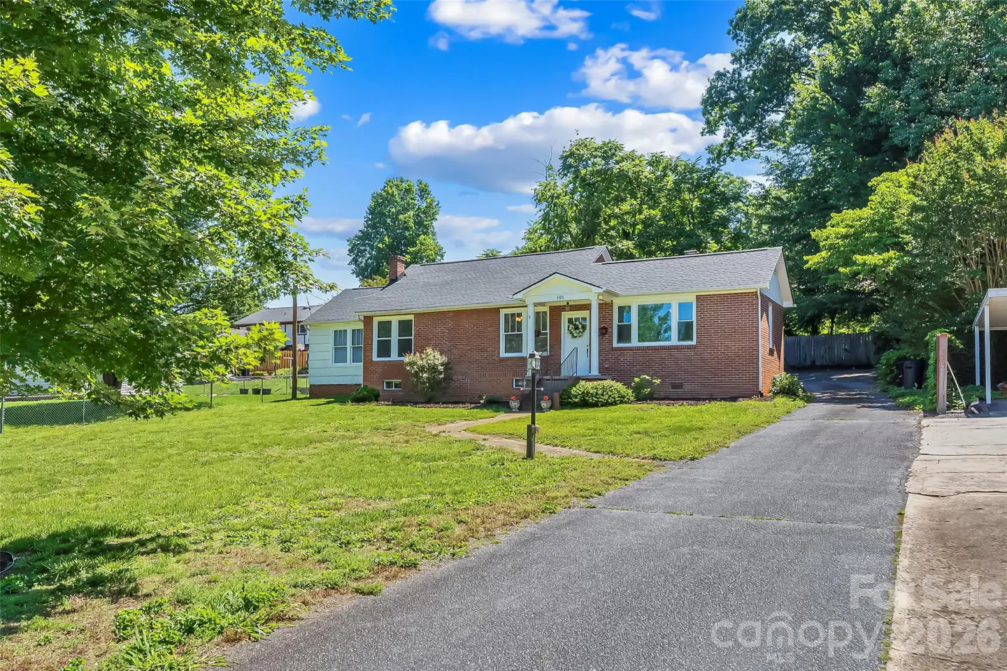Single-story red brick house with a gray roof, white trim, and a long driveway on a sunny day with a green lawn.