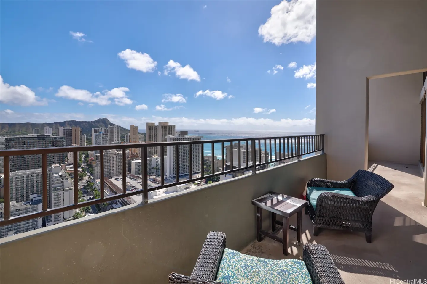 Balcony view of Honolulu, Hawaii. Wicker chairs and a small table sit on the balcony overlooking the city, ocean, and Diamond Head under a blue sky.