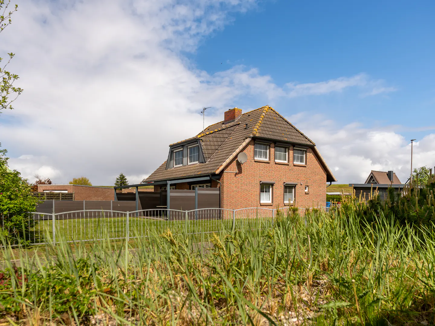 A two-story red brick house with a brown tiled roof, seen from behind a metal fence and tall grass under a blue sky.