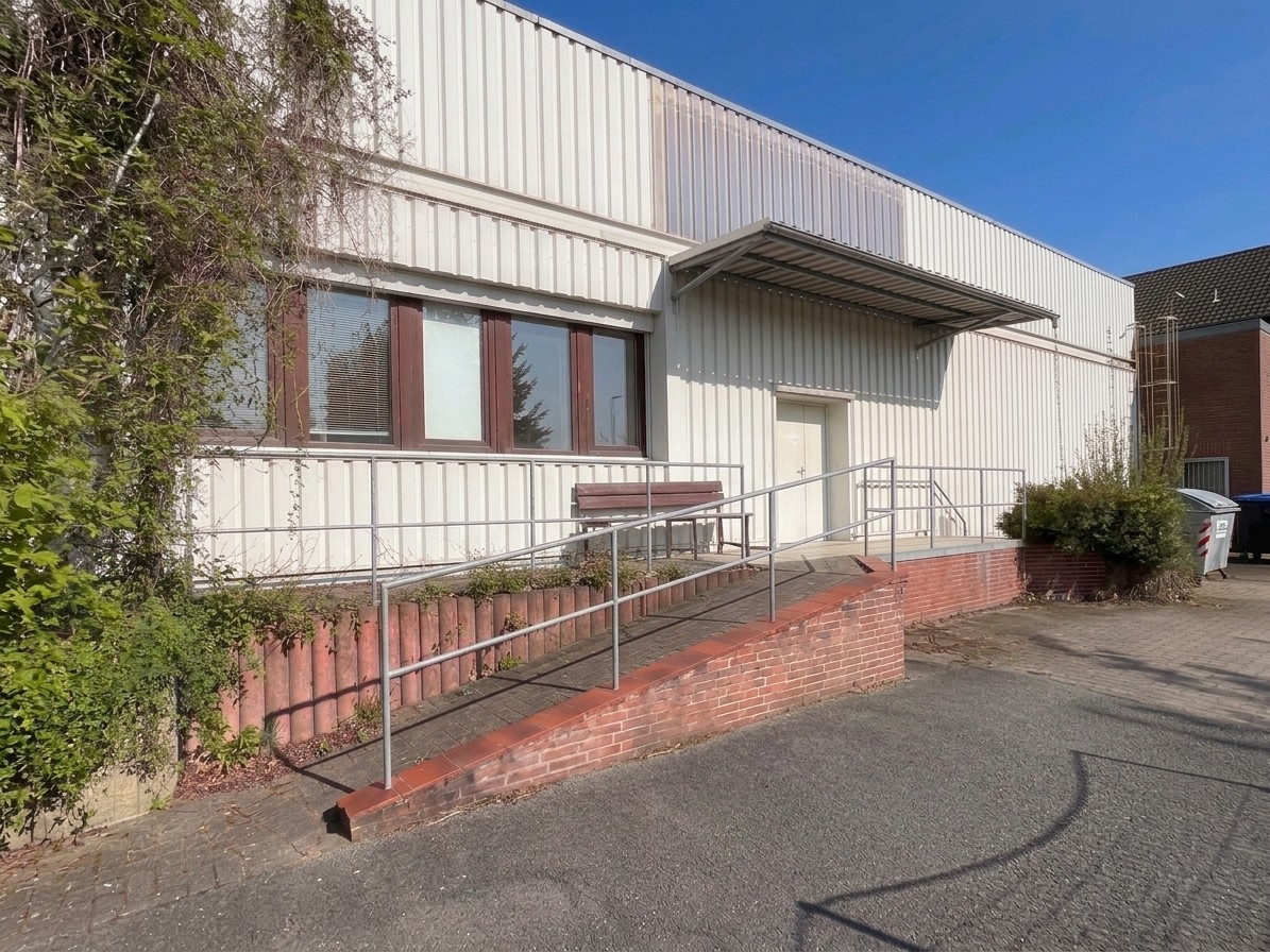 Exterior of a white industrial building with a brick wheelchair ramp, metal railings, and a bench under a canopy. [AI-generated text]