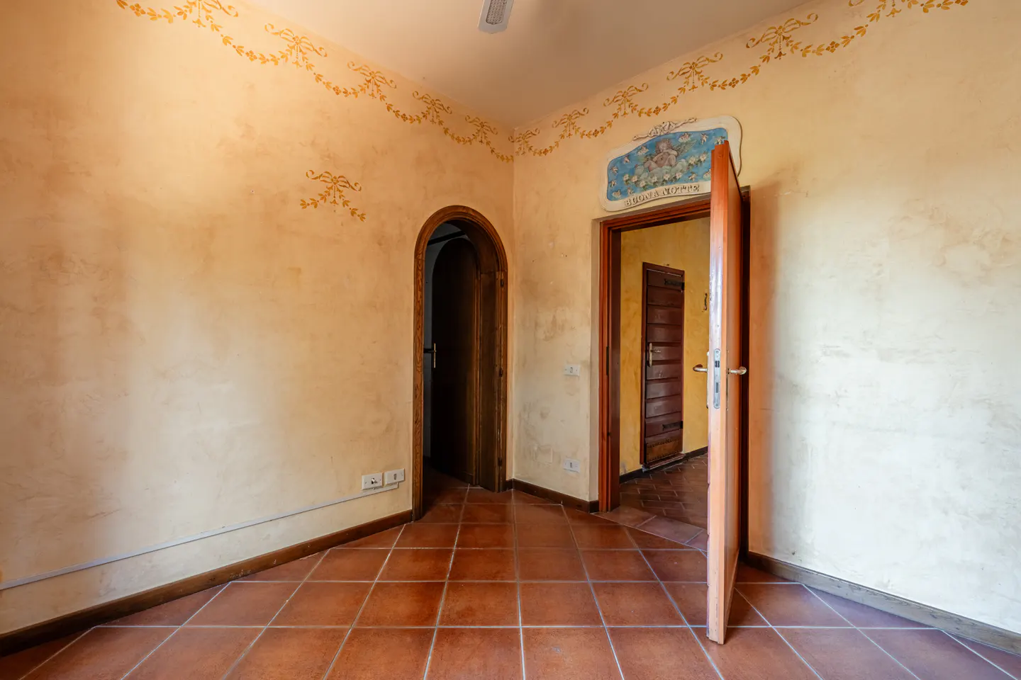 A room with terracotta tile flooring, light yellow walls with gold floral trim, and two open wood doorways.