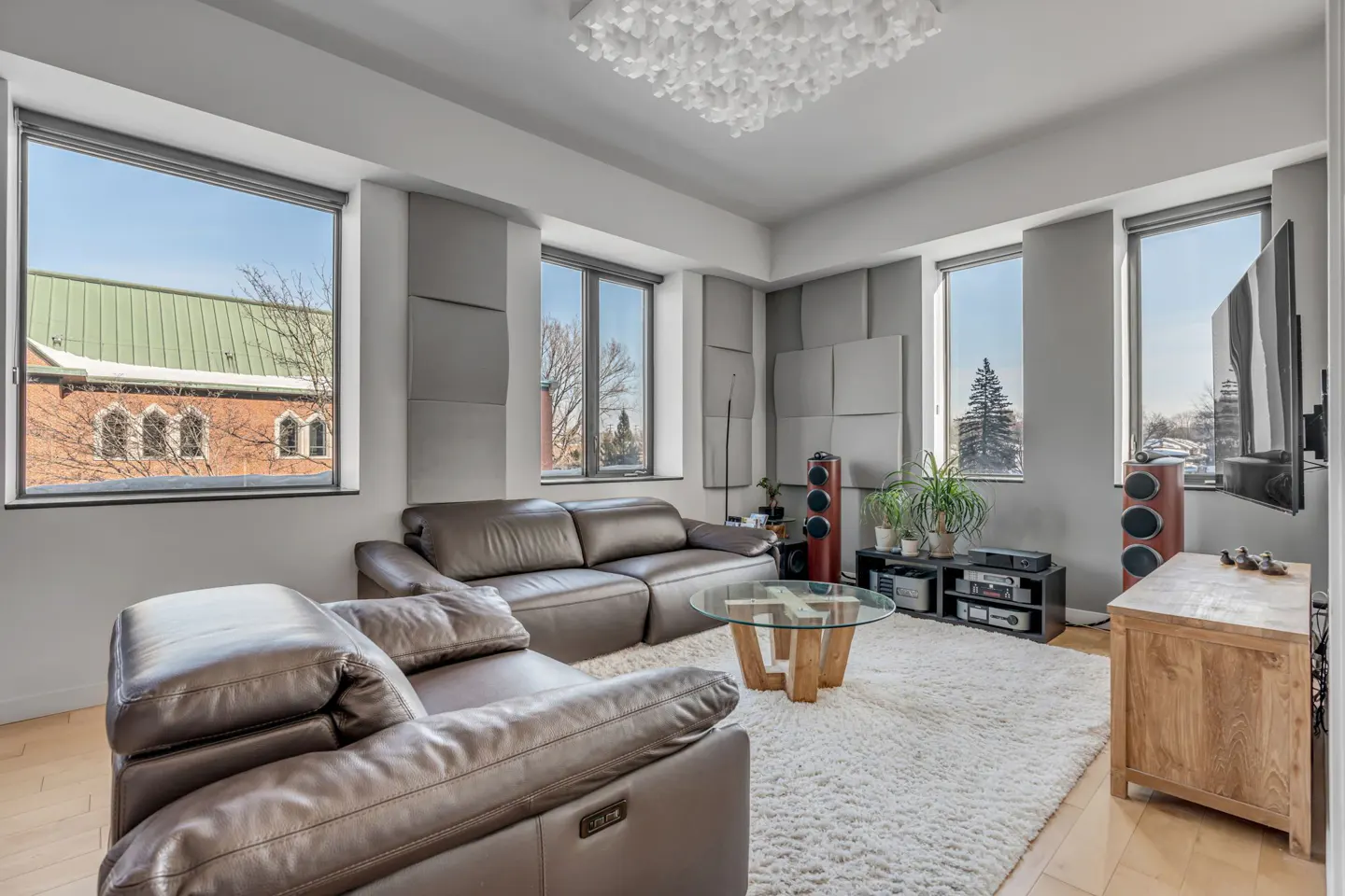 Bright living room with brown leather sofas, a glass table, and a fluffy white rug. Large windows offer city views. Gray soundproofing panels line the walls.