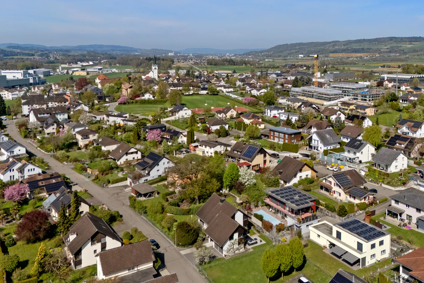 Aerial view of a suburban neighborhood with houses, trees, and a church steeple in the background. Some houses have solar panels.