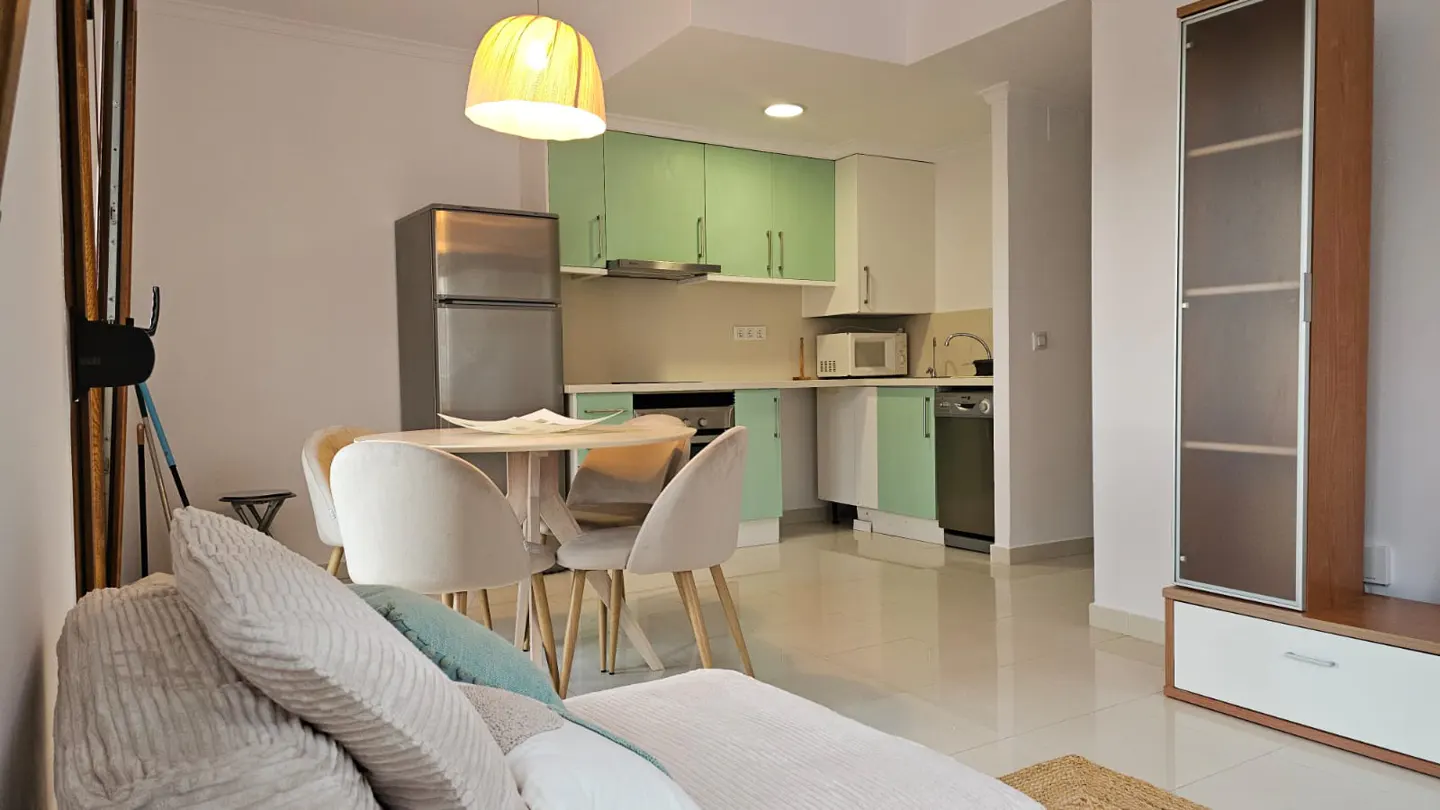 A bright apartment interior with a kitchen, dining table, and sofa. The kitchen has mint green and white cabinets, and stainless steel appliances. A round table with four chairs sits nearby. A sofa with pillows is in the foreground.