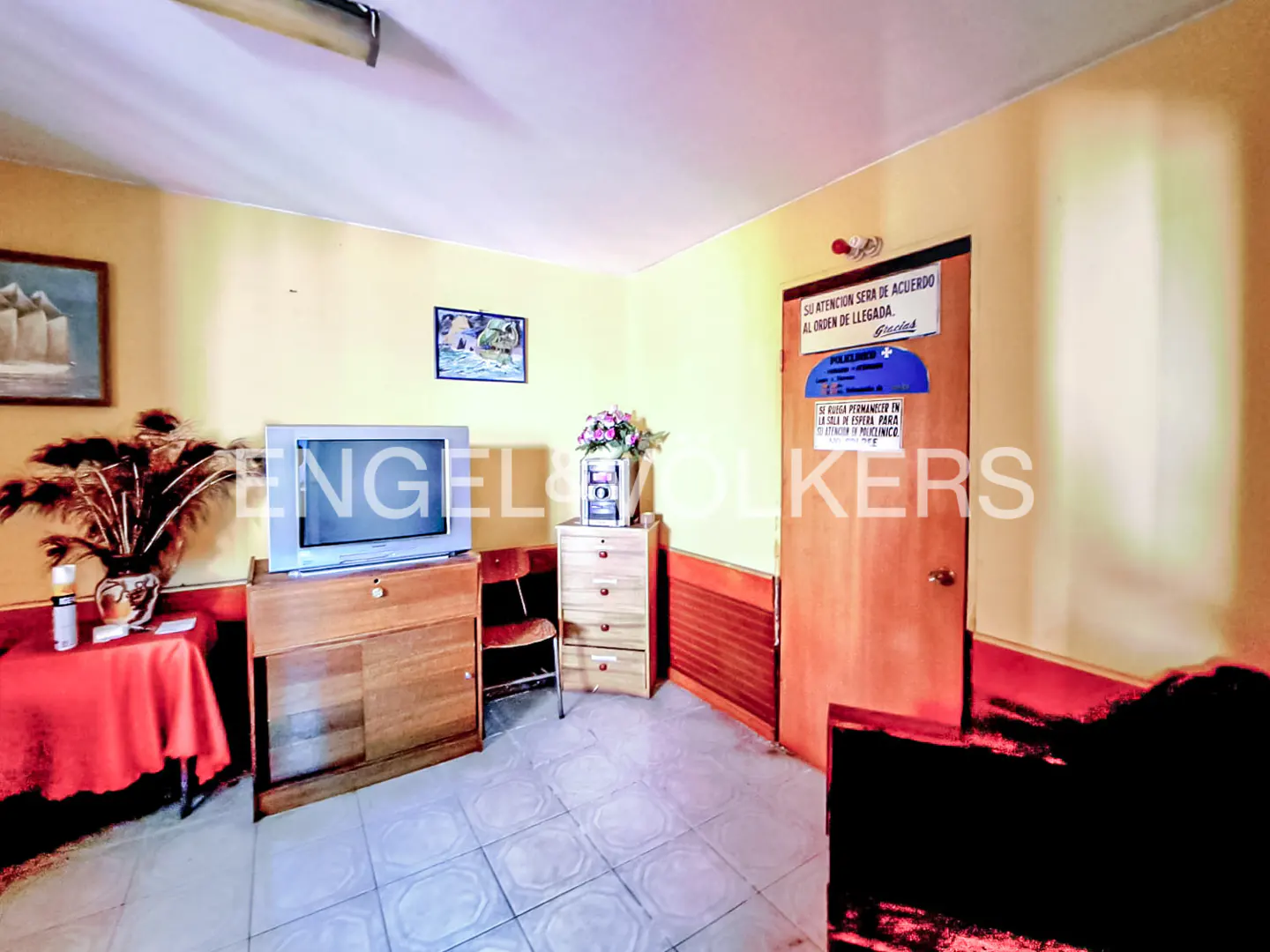 A room with yellow walls, a TV on a wooden cabinet, a red table, and a door with signs.