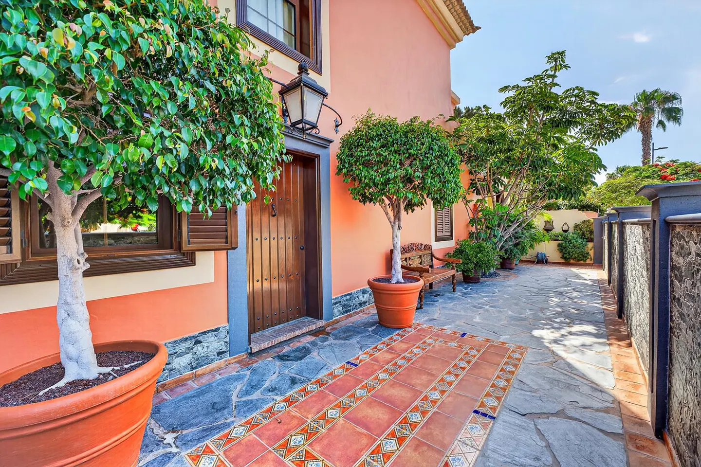 A peach-colored house exterior with a wooden door, potted trees, a decorative tiled walkway, and a stone patio.