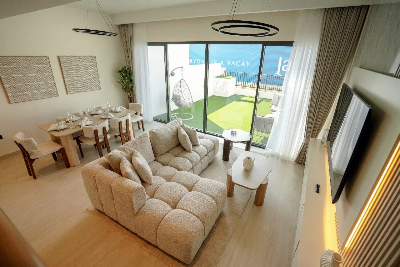Bright living room with a beige sectional sofa, dining table set for six, and a patio with a hanging chair visible through a large window.