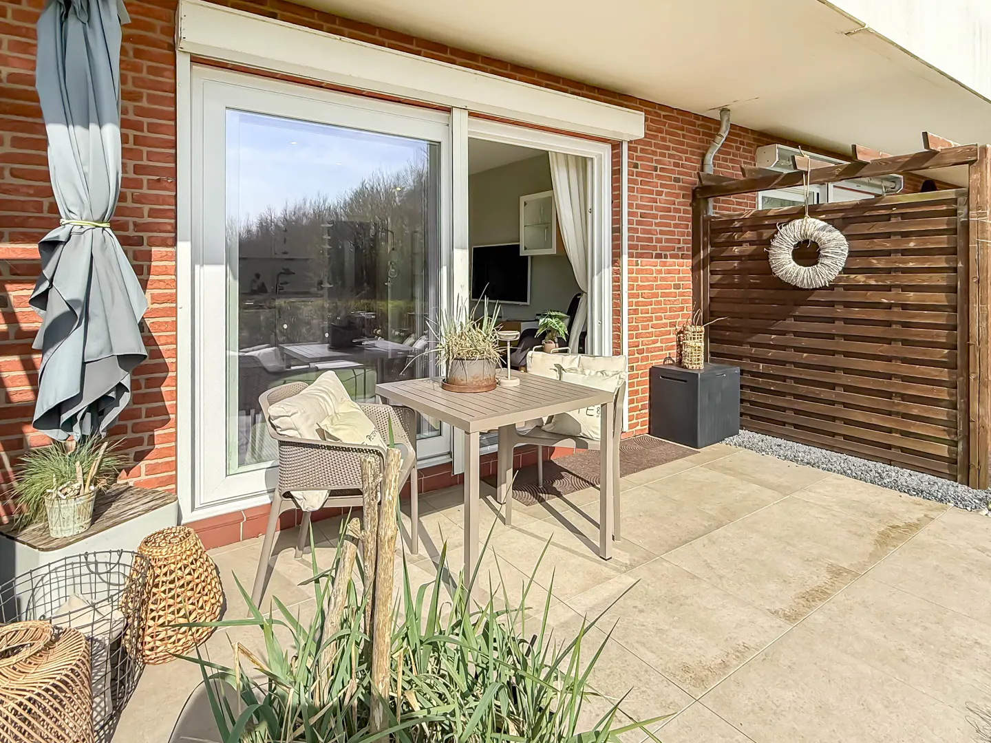 Outdoor patio with a table, chairs, and privacy screen. A rolled-up umbrella is on the brick wall. Sliding glass doors lead to the interior.